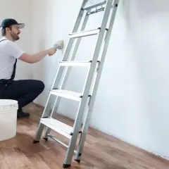 Hombre con brocha y cubo de pintura, listo para limpiar pared antes de pintar. Escalera metálica al lado.