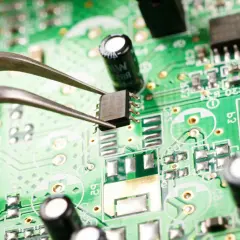 Pęseta trzyma układ scalony na zielonej płytce drukowanej. Widać elementy elektroniczne, kondensatory i ścieżki. Zbliżenie ujawnia potencjalne oznaki korozji elektrochemicznej.