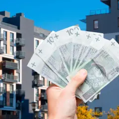 Dłoń trzyma banknoty 100 zł na tle nowoczesnych budynków mieszkalnych. Symbolizuje to podatek od sprzedaży nieruchomości.
