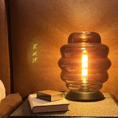 Una lámpara de mesa de cristal ahumado ilumina un rincón acogedor junto a una cama con almohada amarilla.