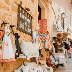 Bunte Stoffe und Kissen hängen vor einem Geschäft am alcudia markt. Menschen schlendern die Gasse entlang.