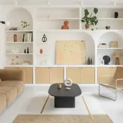 Salon épuré avec canapé beige, fauteuil en toile, table basse noire et étagères murales remplies d'objets décoratifs. Le tout baigne dans une douce lumière, rehaussée par la **couleur blanc cassé** des murs et des étagères.