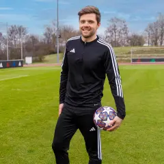 Uśmiechnięty mężczyzna w stroju sportowym trzyma piłkę na boisku. To jego pierwszy staż trenerski, pełen nowych wyzwań i nauki.