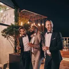 Trzech mężczyzn w garniturach trzymających drinki, śmiejących się na tle nocnego przyjęcia.