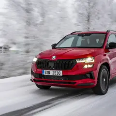 Czerwony crossover 4x4 pokonuje ośnieżoną drogę w zimowej scenerii, z domkiem w tle.