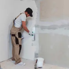 Operaio con tuta beige e cappellino nero mentre impara come intonacare un muro, usando una spatola. Scala e secchio a terra.