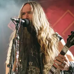 Zakk Wylde, wokalista i gitarzysta, z długimi blond włosami i naszyjnikami, gra na gitarze podczas koncertu.