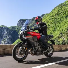 Un motociclista su una Yamaha Tracer rossa affronta una curva su una strada panoramica, godendosi una gita in moto tra montagne verdi e cielo azzurro.