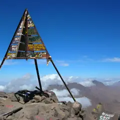 Sommet du mont Toubkal, altitude impressionnante. Le cairn triangulaire, couvert de graffitis, se dresse fièrement sur un pic rocheux, dominant un paysage montagneux spectaculaire sous un ciel bleu éclatant.