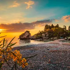 Tramonto dorato sulle spiagge di Giardini Naxos Taormina, con rocce e vegetazione lussureggiante.
