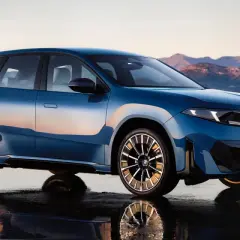 La BMW Neue Klasse blu metallizzato, con riflessi del tramonto, si staglia su uno sfondo montuoso.