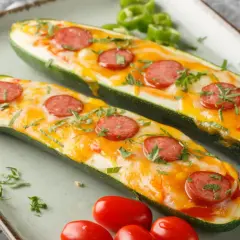 Dwa pyszne łódeczki z cukinii faszerowane warzywami i mozzarellą, posypane natką pietruszki i kawałkami kiełbasy.