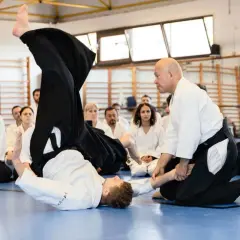 Två personer i vit gi utför en kastteknik inom kampsporten aikido. Åskådare sitter på golvet och observerar.