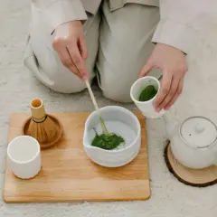 Hände bereiten Matcha zu. Ein Bambusbesen und eine Tasse stehen bereit. Dieser Matcha macht wach!