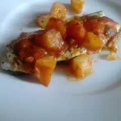 Un delicioso jargo pescado cubierto con salsa de tomate y trozos de patata, servido en un plato blanco.