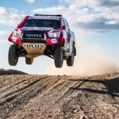Toyota Hilux w locie podczas rajdu Dakar. Widzowie obserwują zmagania na pustynnym terenie.