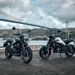 Dwa motocykle Husqvarna Svartpilen 250 stoją nad wodą, pod mostem wiszącym, w malowniczym, górzystym krajobrazie.