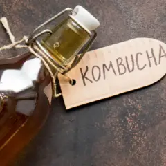En flaska med en mörk, bubblig kombucha dryck. En brun etikett med texten "Kombucha" hänger på flaskhalsen.