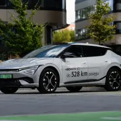 Biały Kia EV6 zaparkowany przed nowoczesnym budynkiem, z napisem "#GOelectric do 528 km zasięgu".