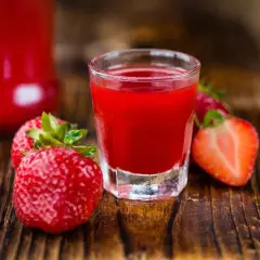 A shot glass filled with vibrant red nalewka z truskawek, surrounded by fresh strawberries on a wooden surface.