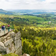 Widok z platformy na skalnym urwisku na malownicze **Góry Stołowe**. Grupa turystów podziwia rozległą panoramę lasów i zielonych wzgórz.