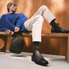 Mężczyzna w niebieskim swetrze i beżowych chinosach prezentuje stylowe buty do chinosów. Elegancka torba na ramię dopełnia całość.
