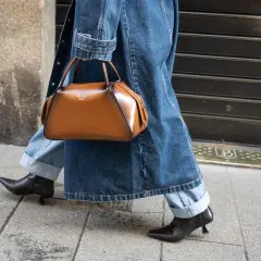 Une femme élégante marche dans la rue, portant un jean retroussé, une veste en jean et un sac à main marron. Le style "comment retrousser son jean femme" est parfait pour cette tenue.