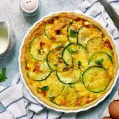 Pyszna tarta z cukinią, zapieczona ze złotym serem i ziołami. Obok dzbanuszek mleka i jajka.