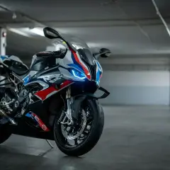 Sportowy motocykl BMW XR w biało-niebiesko-czerwonych barwach, z włączonymi światłami LED, stoi na betonowej posadzce w garażu.