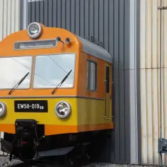 Pomarańczowo-żółty pociąg EW58-019sa stoi przed halą.