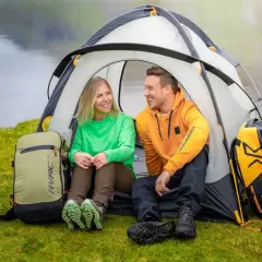 Ett glatt par njuter av att campa i skogen vid en sjö. De sitter i sitt tält med ryggsäckar bredvid.