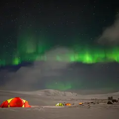 Nocne niebo nad Norwegią rozświetla zorza polarna. Kilka kolorowych namiotów rozbitych na śniegu tworzy przytulne schronienie pod tym niezwykłym zjawiskiem.