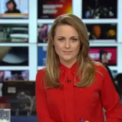 Marta Kłos, której wiek nie jest widoczny, w czerwonej bluzce z kokardą, na tle ekranów informacyjnych.