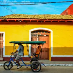 Un hombre pedalea un taxi bicitaxi frente a una casa amarilla vibrante, un recuerdo de mi viaje a Cuba.