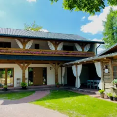 Drewniane domki i budynek z werandą, ozdobione kwiatami, na tle zieleni i błękitnego nieba.