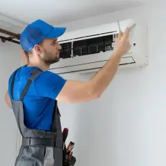 Technik w niebieskiej czapce i ogrodniczkach naprawia klimatyzator.