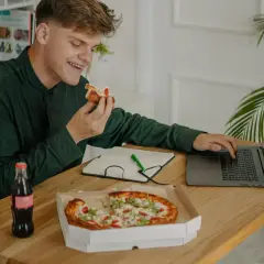 Młody mężczyzna je pizzę, pracując przy laptopie. Obok butelka napoju i notatnik. Dba o dietę, pamiętając o insulinie aspart.