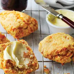 Trois scones américains dorés, dont un coupé en deux et garni de beurre, sur une grille. Un pot de confiture et du beurre à tartiner sont à proximité.