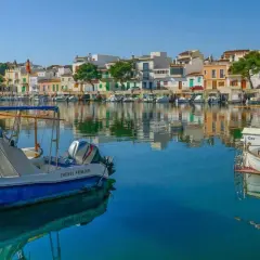 Boote im Hafen von Portocolom, Mallorca, spiegeln sich im ruhigen, blauen Wasser vor bunten Häusern.