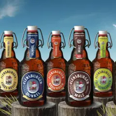 Flensburger Pils Bier Marken am Strand. Eine Möwe fliegt über das Meer, während die Flaschen mit Bügelverschluss im Gras stehen.