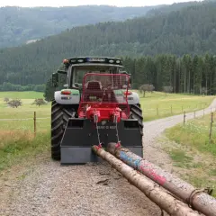 Traktor mit angehängter seilwinde forst zieht zwei Baumstämme über einen Schotterweg, umgeben von grünen Wiesen und einem dichten Wald.