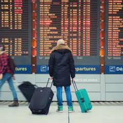 Tablica odlotów na lotnisku z podróżnymi ciągnącymi walizki.