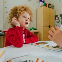 Chłopiec z kręconymi włosami, w czerwonym sweterku, uczy się z kart obrazkowych. Zastanawiamy się, ile godzin wspomagania dla ucznia z autyzmem jest potrzebne.