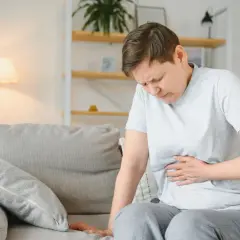 Kobieta siedzi na kanapie, trzymając się za brzuch. Jej twarz wyraża ból, co może sugerować przewlekłe zapalenie trzustki.