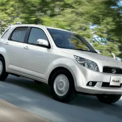 Biały Toyota Rush pędzi po drodze, otoczony zielenią drzew.