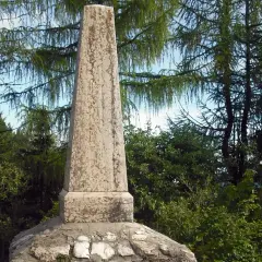 Obelisco in pietra sul **monte Zugna**, circondato da alberi di larice e vegetazione rigogliosa.