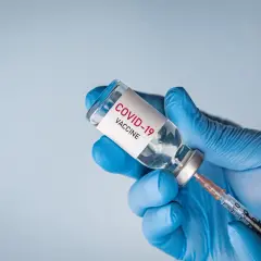 Ręka w niebieskiej rękawiczce trzyma fiolkę z napisem "COVID-19 VACCINE" i strzykawkę, gotowa do szczepienia.