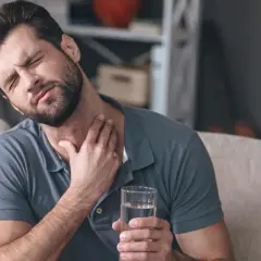 Mężczyzna z zamkniętymi oczami trzyma szklankę wody, czując nieprzyjemne drapanie w gardle.