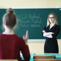 Nauczycielka w okularach stoi przed tablicą z zapisanymi czasami gramatycznymi.