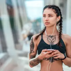Kobieta z warkoczami i słuchawkami na uszach, trzymająca telefon. Widoczny delikatny tatuaż między piersiami w kształcie kota.